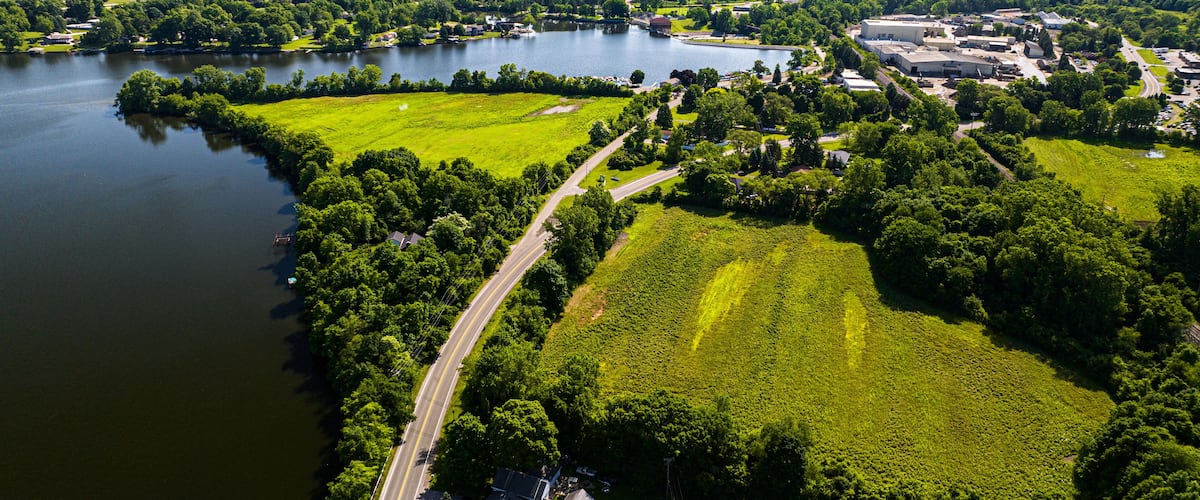 An aerial view of a lakeside area with a road, green fields, and buildings.