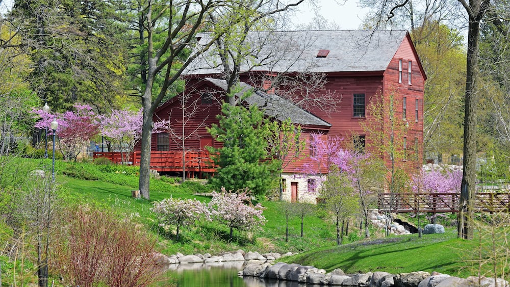 Une maison rouge entourée d’un étang, d’arbres et de fleurs en pleine floraison.