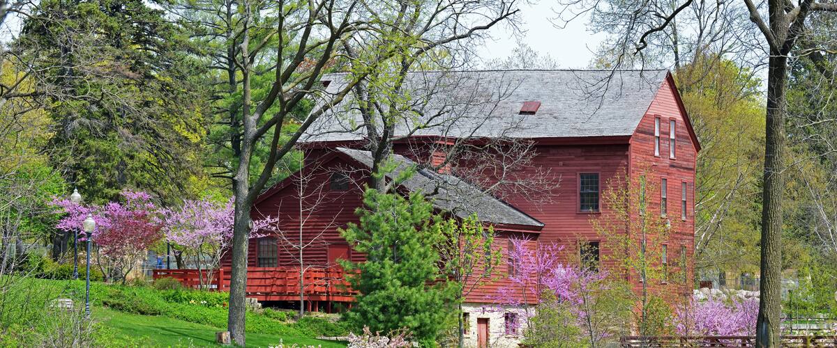 A red house surrounded by a pond, trees, and blooming flowers.