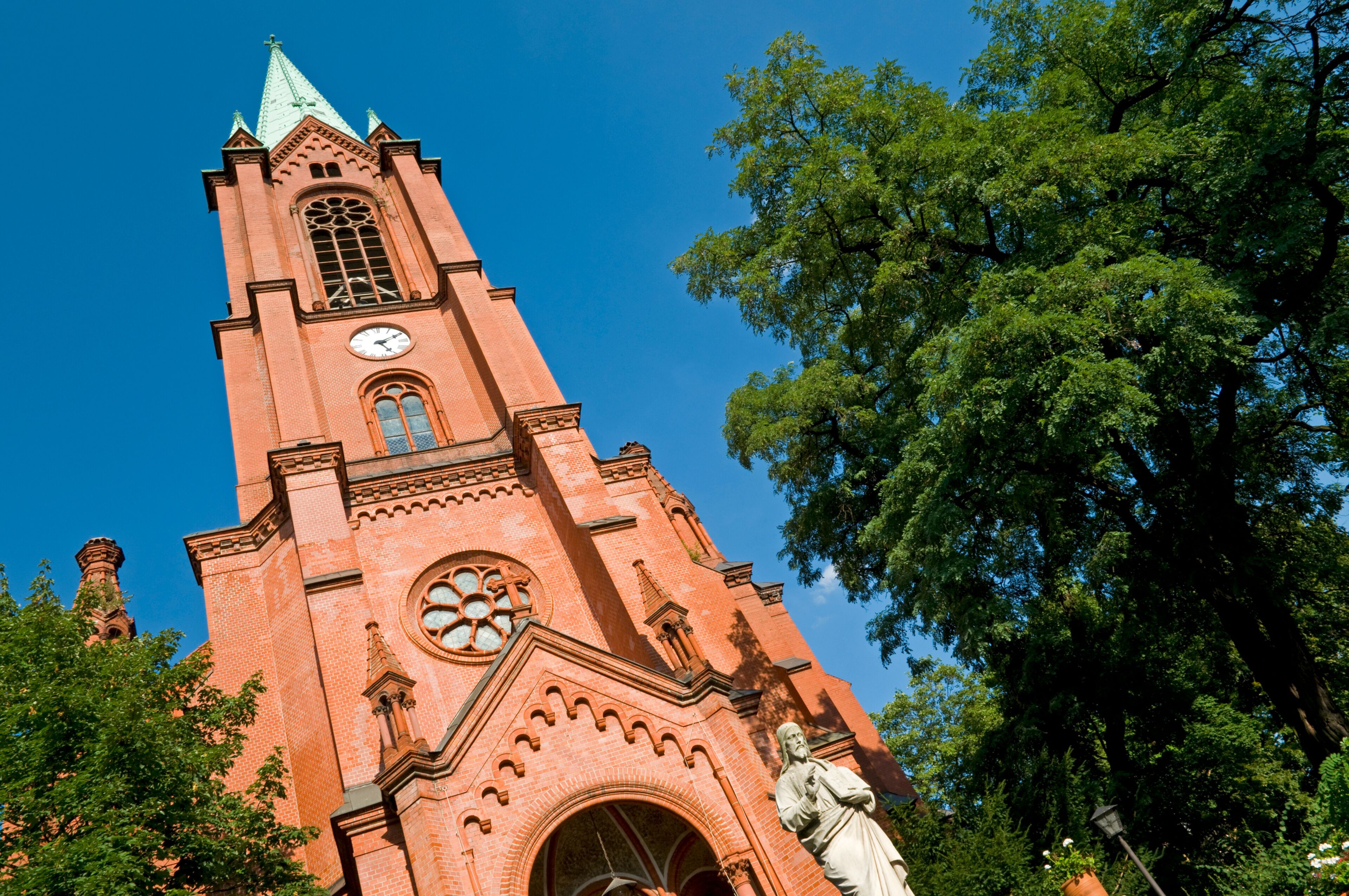 A tall red brick church with a clock tower, a large rose window, and a statue in front.