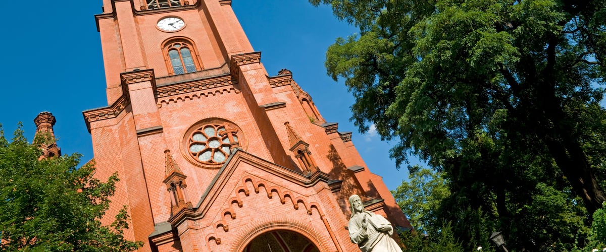 A tall red brick church with a clock tower, a large rose window, and a statue in front.