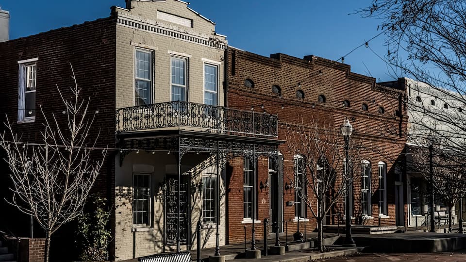 A row of historic brick buildings with a covered balcony and a bench on the sidewalk.