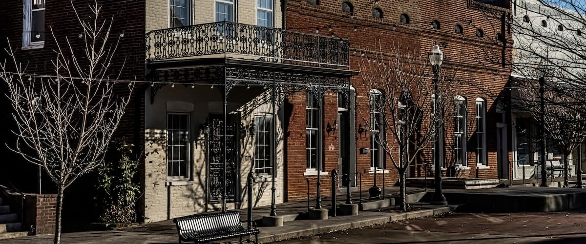 A row of historic brick buildings with a covered balcony and a bench on the sidewalk.