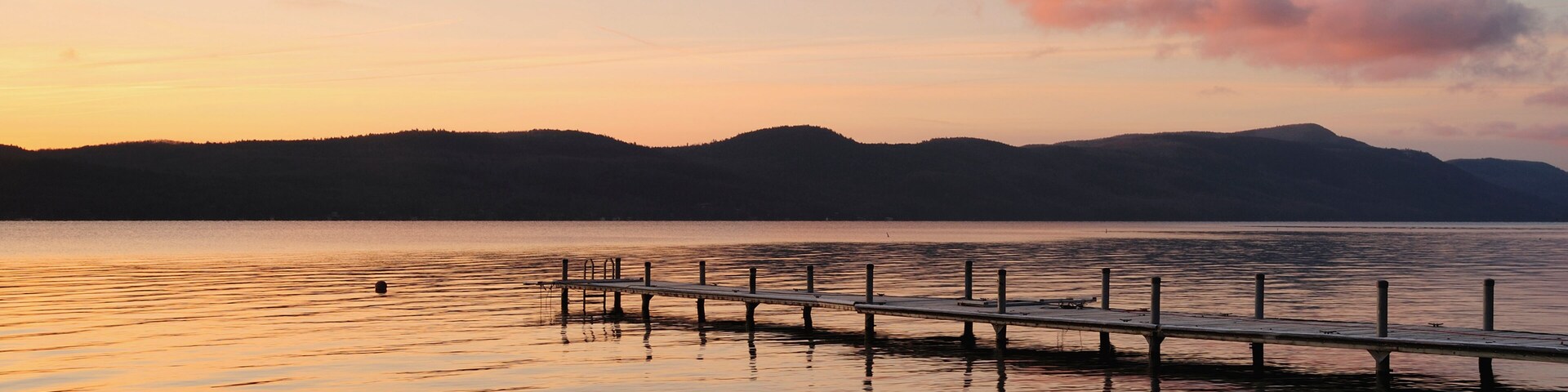 Un lago tranquilo al atardecer con un muelle de madera que se adentra en el agua, rodeado de montañas.