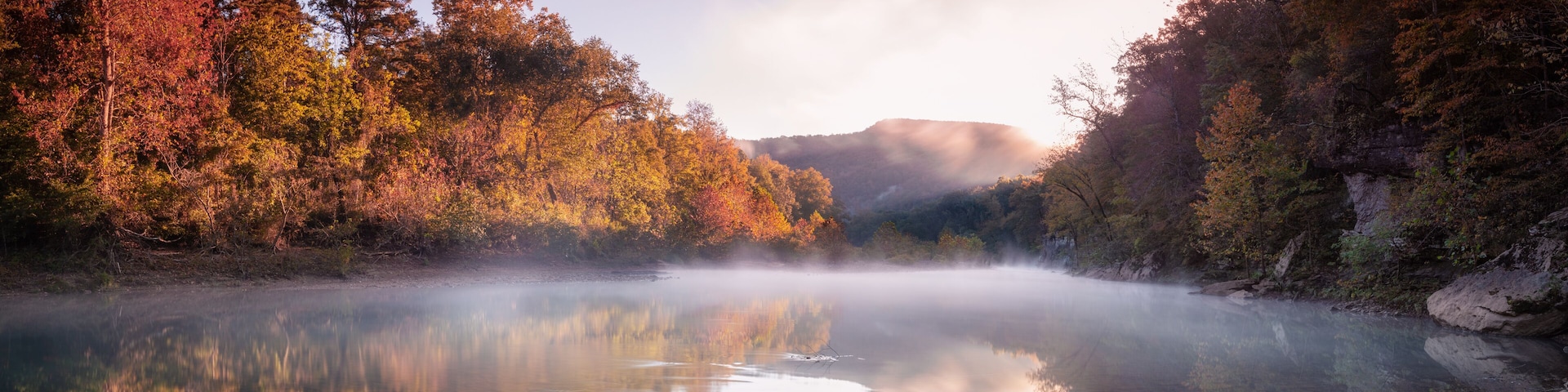 A calm river surrounded by autumn-colored trees and a misty mountain backdrop.
