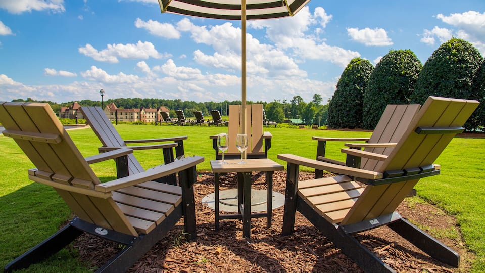 a gorgeous summer landscape at the vineyard with a brown wooden tables and chairs in a circle around a brown and white umbrella with wine glass on the table surrounded lush green trees and grass