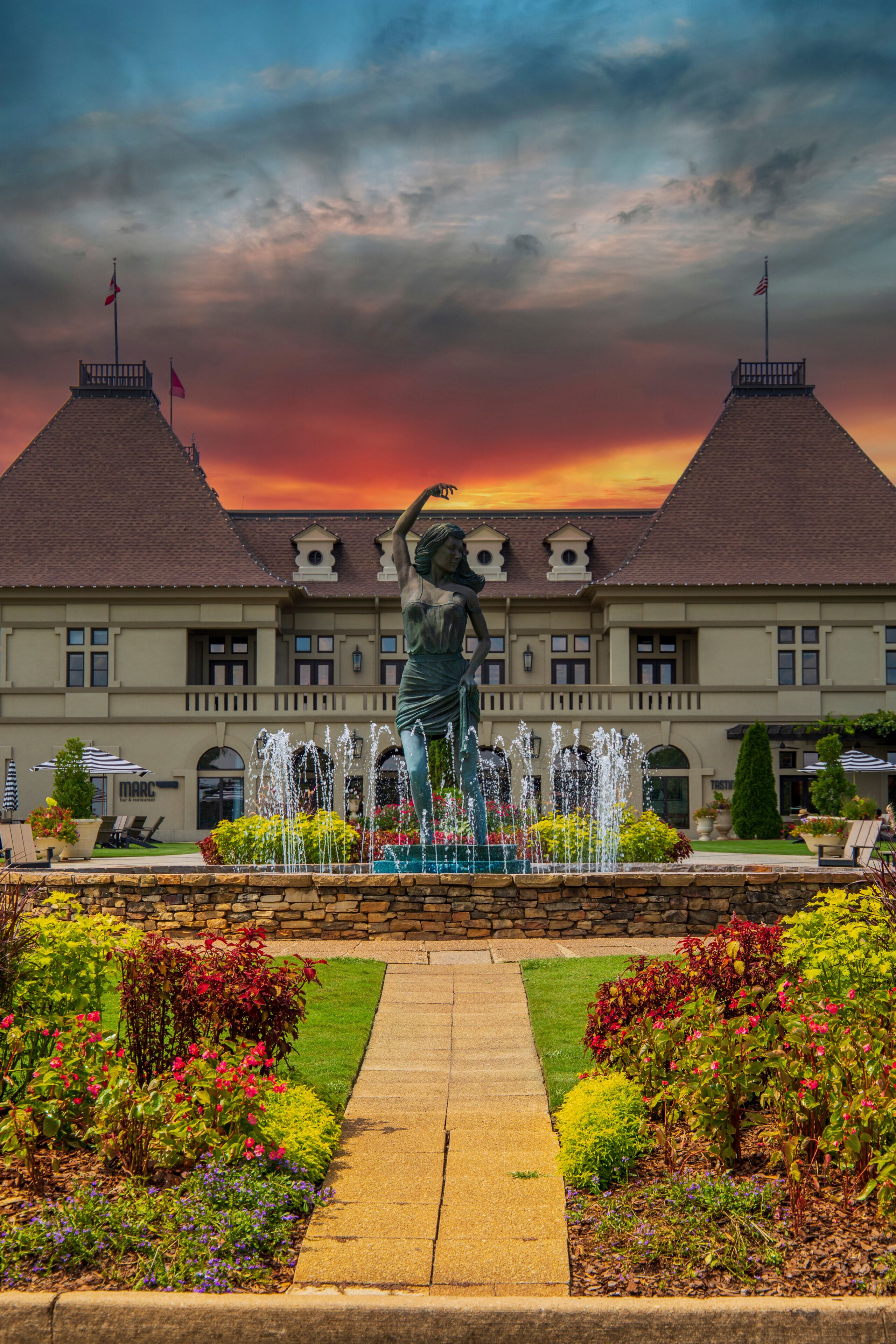 a gorgeous summer landscape with a water fountain with statue of a woman in the center surrounded by colorful flowers and lush green trees, grass and plants with red sky and powerful clouds	