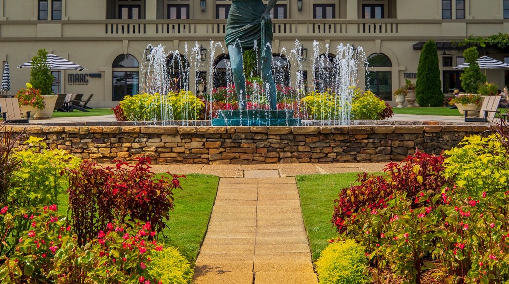 a gorgeous summer landscape with a water fountain with statue of a woman in the center surrounded by colorful flowers and lush green trees, grass and plants with red sky and powerful clouds