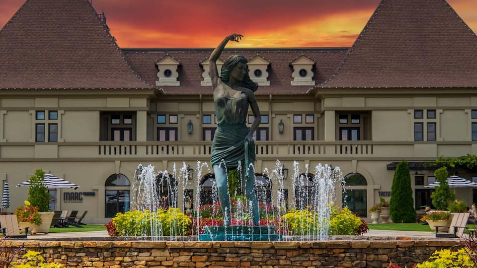 a gorgeous summer landscape with a water fountain with statue of a woman in the center surrounded by colorful flowers and lush green trees, grass and plants with red sky and powerful clouds