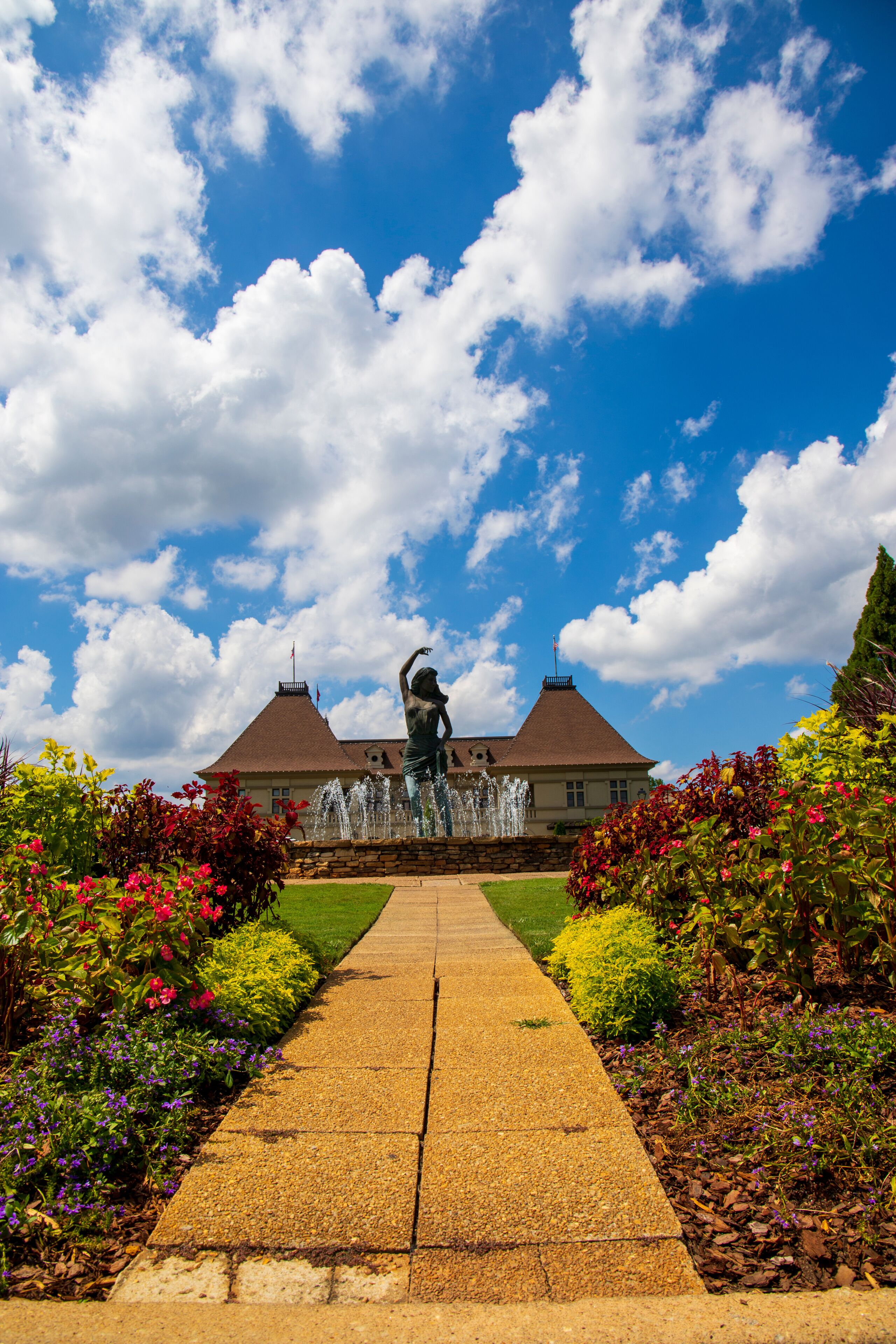 a gorgeous summer landscape with a water fountain with statue of a woman in the center surrounded by colorful flowers and lush green trees, grass and plants with blue sky and powerful clouds