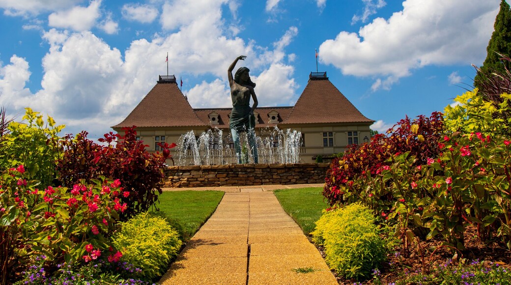 a gorgeous summer landscape with a water fountain with statue of a woman in the center surrounded by colorful flowers and lush green trees, grass and plants with blue sky and powerful clouds