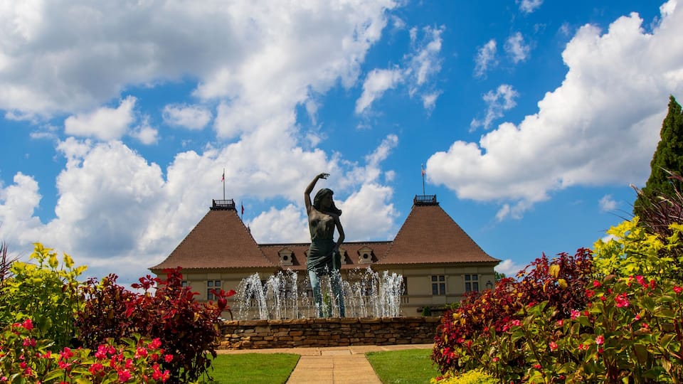 a gorgeous summer landscape with a water fountain with statue of a woman in the center surrounded by colorful flowers and lush green trees, grass and plants with blue sky and powerful clouds