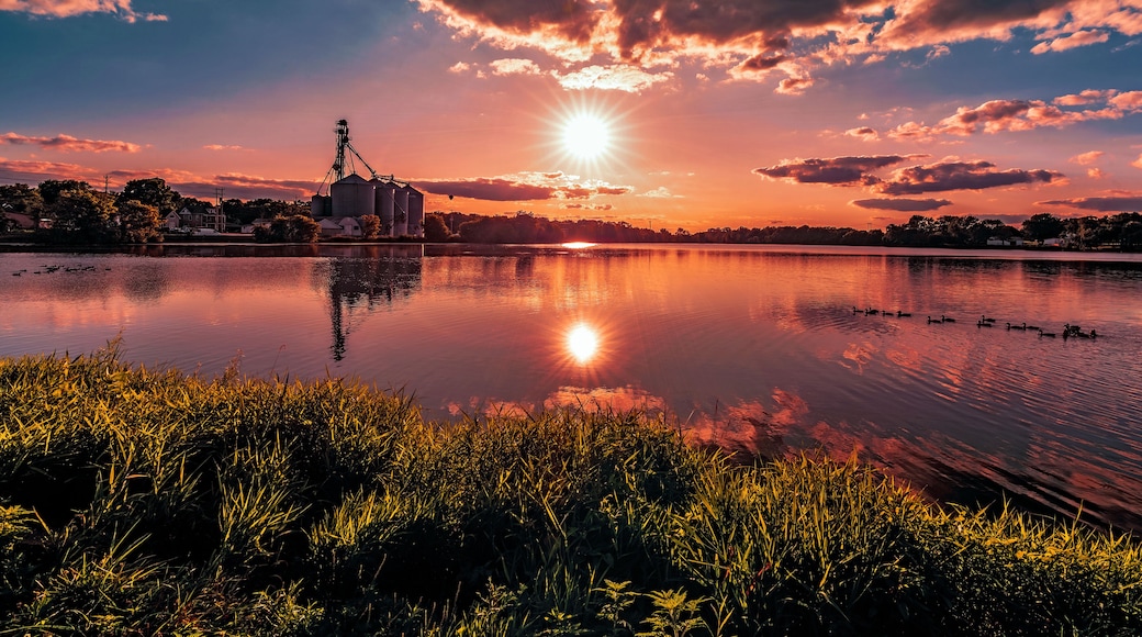 On the grassy shore of a rural southern Wisconsin lake with a view of the sun setting over the silhouettes in the distance at sunset in Burlington, Wisconsin USA.