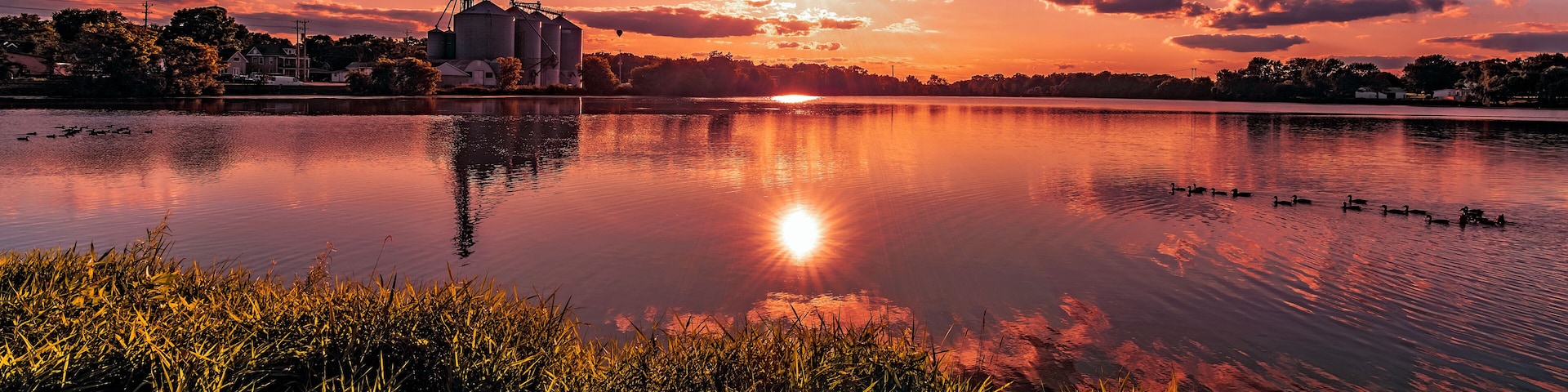 On the grassy shore of a rural southern Wisconsin lake with a view of the sun setting over the silhouettes in the distance at sunset in Burlington, Wisconsin USA.