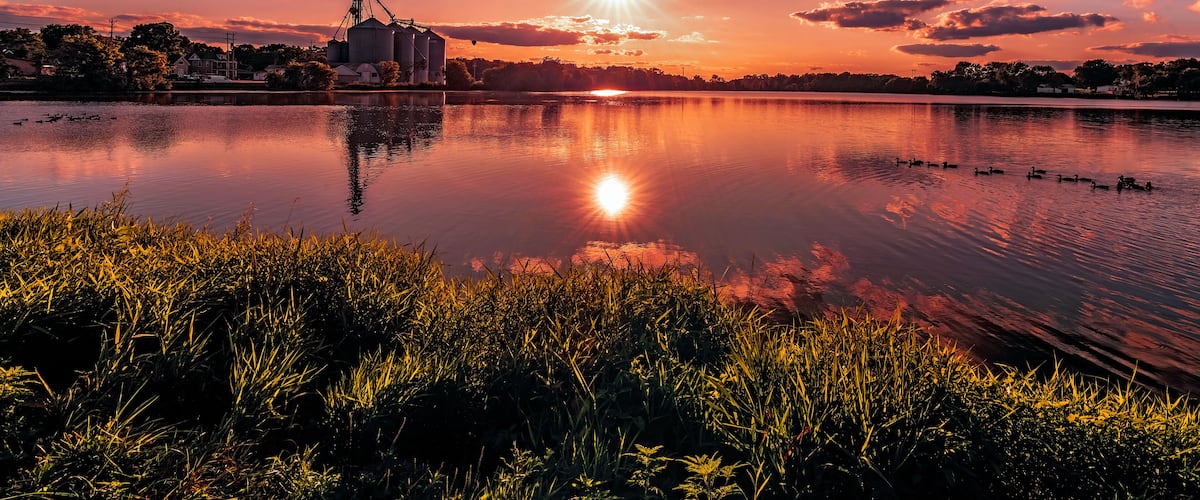 A sunset over a calm lake with industrial buildings in the background.