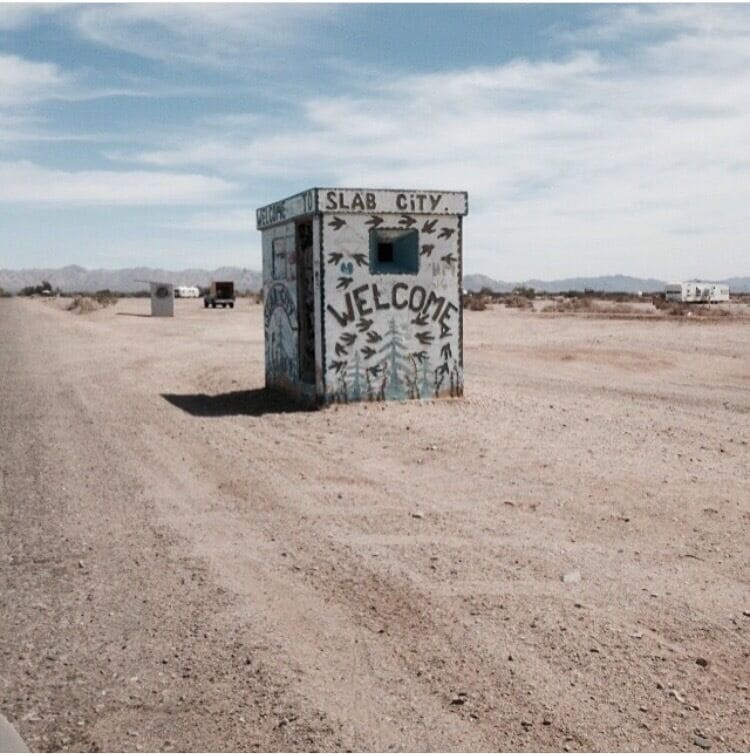 A signpost labeled 'Slab City' with 'Welcome' written on it, set against a desert landscape.
