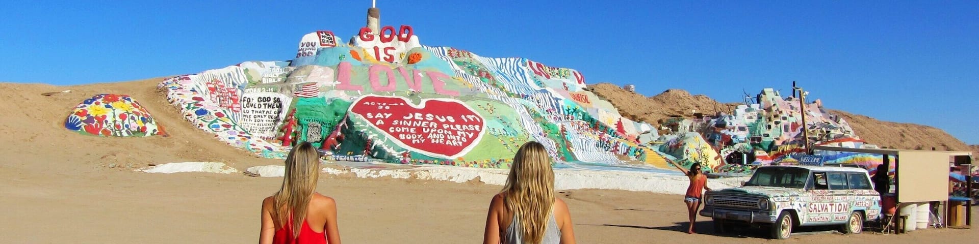 Two women walking on a sandy beach with colorful graffiti art in the background.