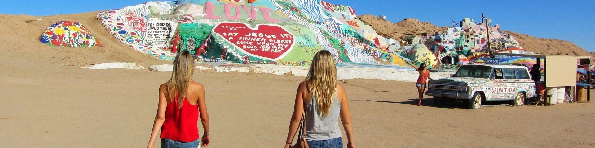 Dos mujeres caminando sobre una playa arenosa con grafitis coloridos al fondo.