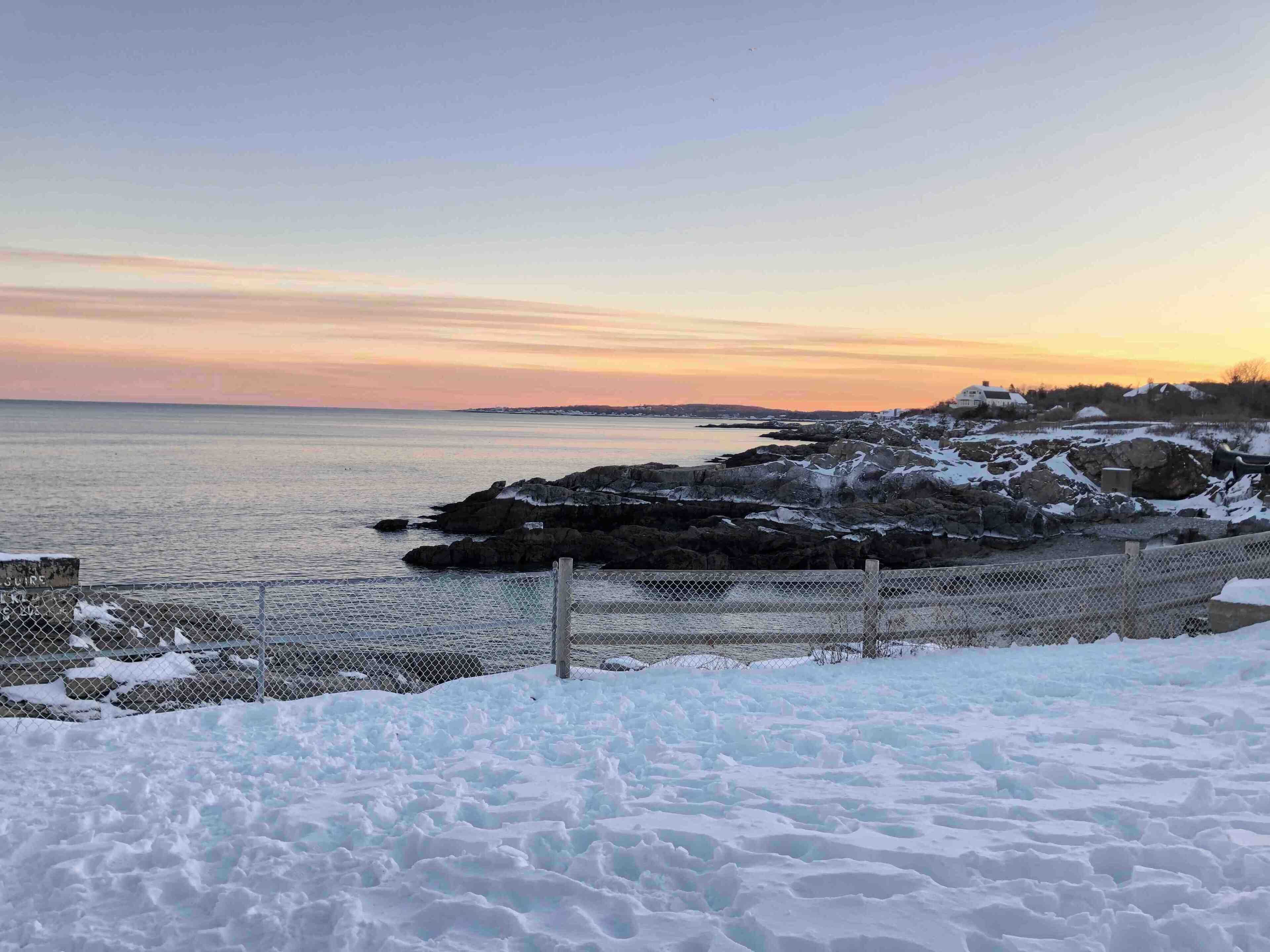 A coastal landscape with snow-covered rocks and a fence at the shoreline.