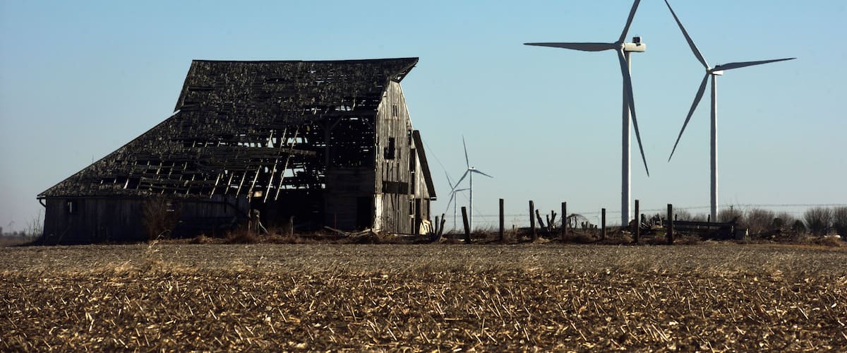 A dilapidated barn, wind turbines, and a field of harvested crops.