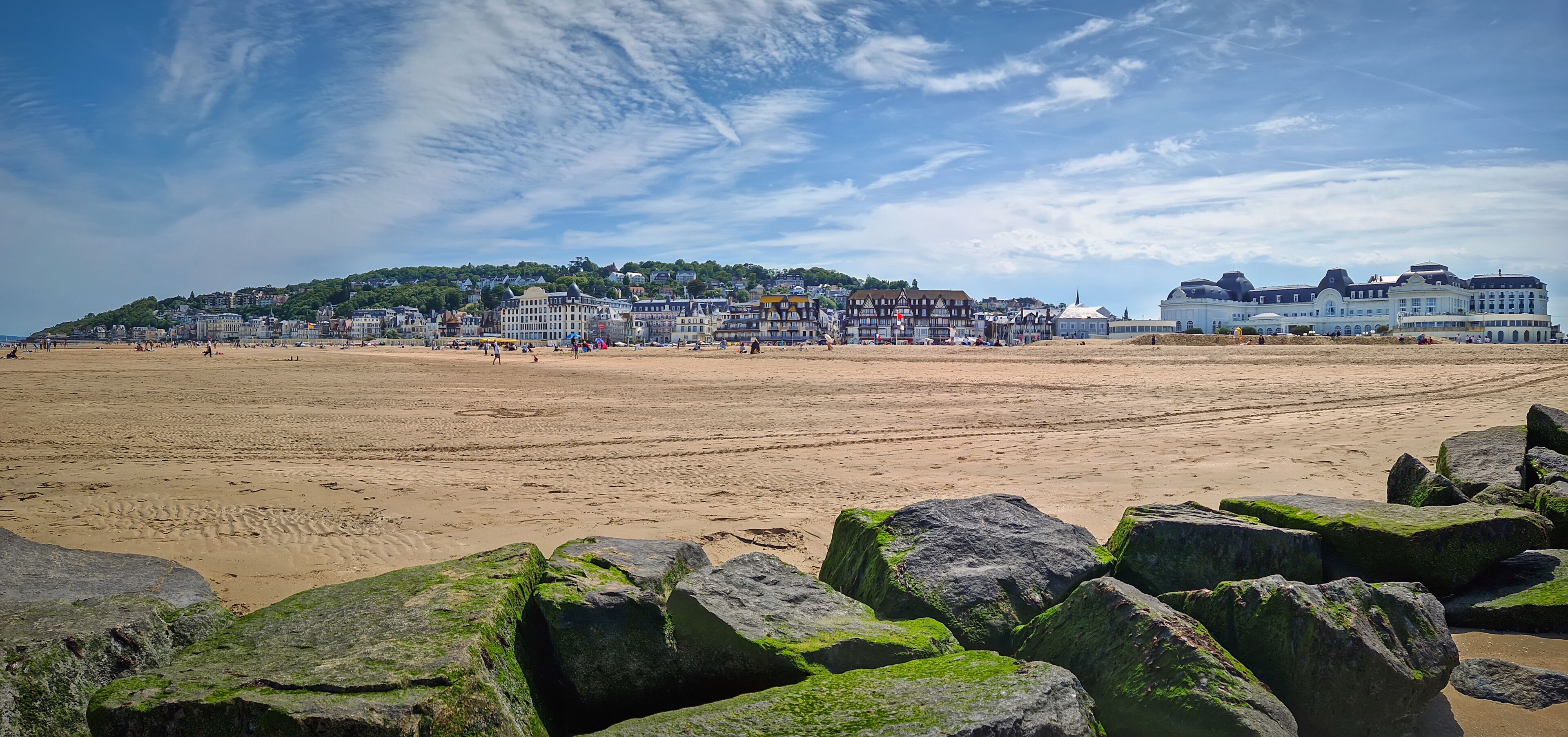 A beach with large rocks in the foreground, a sandy shore, and a town with historic buildings in the background.
