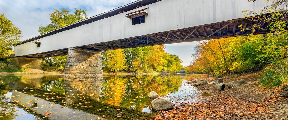 A covered bridge over a calm river with autumn leaves on the ground.