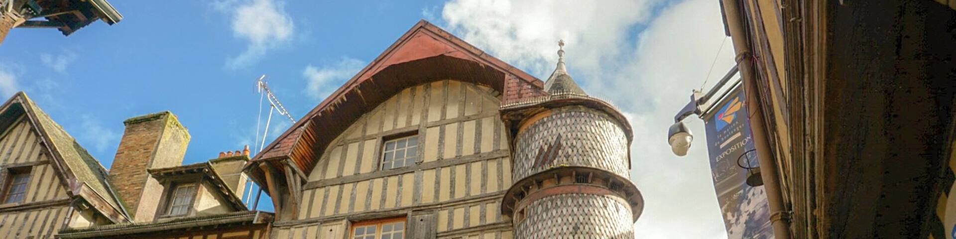 A historic building with timber framing, a cylindrical tower, and a church in the background.