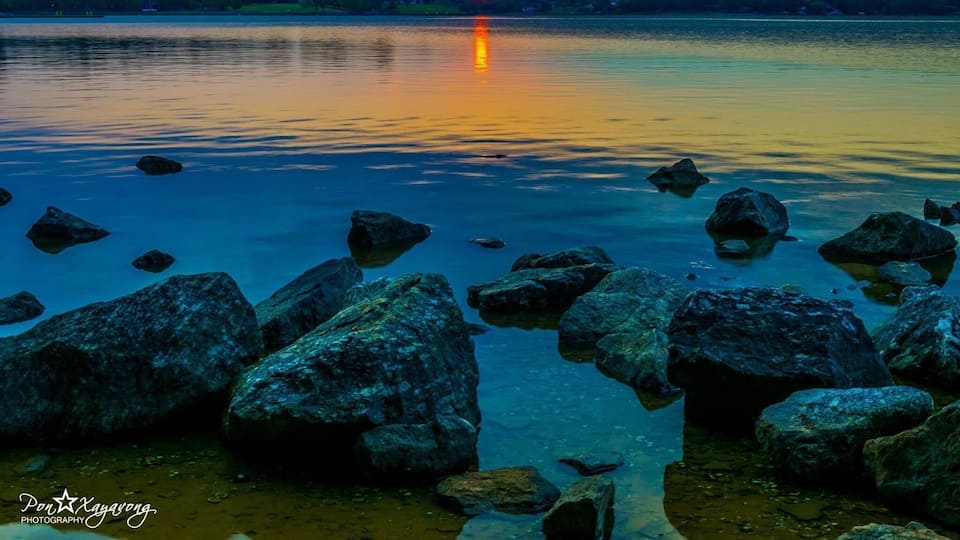 A sunset over a calm lake with large rocks in the foreground.