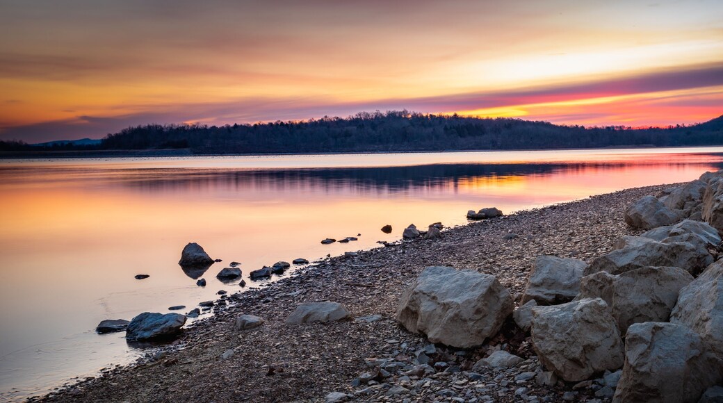 A calm lake at sunset with a rocky shoreline and a forested hill in the background.