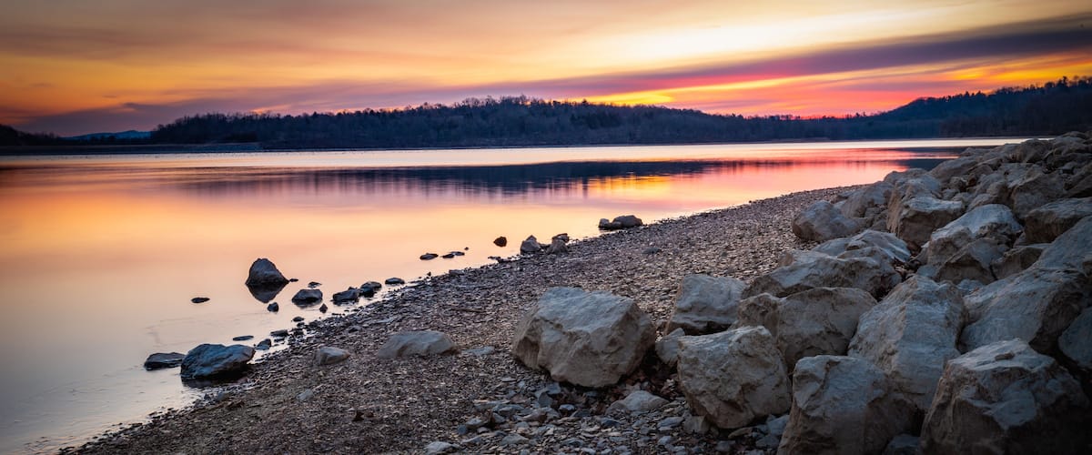 Un lago tranquilo al atardecer con una costa rocosa y una colina boscosa al fondo.