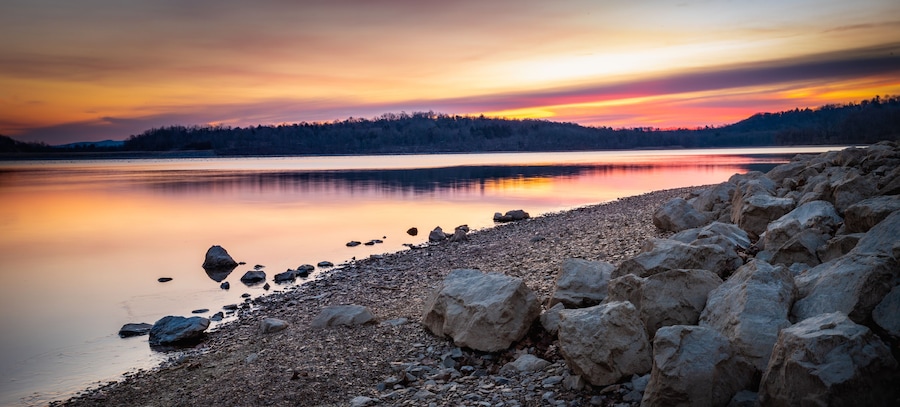 Un lago tranquilo al atardecer con una costa rocosa y una colina boscosa al fondo.