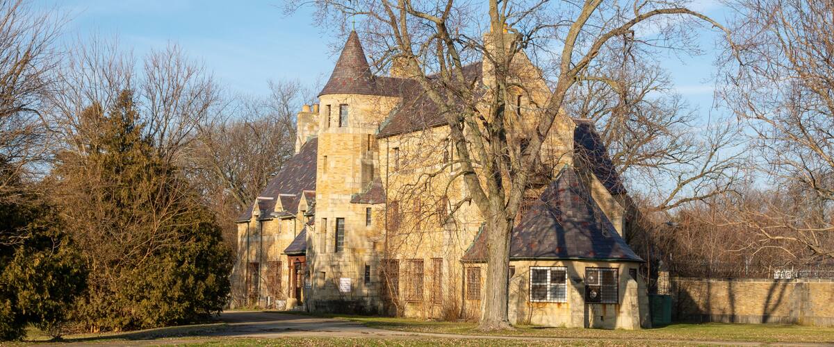 A historic stone building with a conical roof, surrounded by trees and a grassy area.