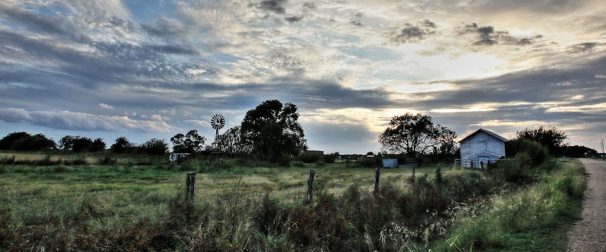 A rural landscape with a windmill, a barn, and a dirt road under a cloudy sky.