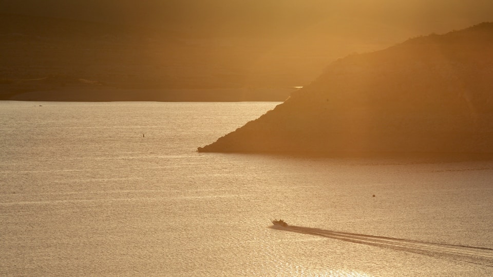 A calm sea with a boat creating a wake, a steep cliff, and the sun setting in the background.