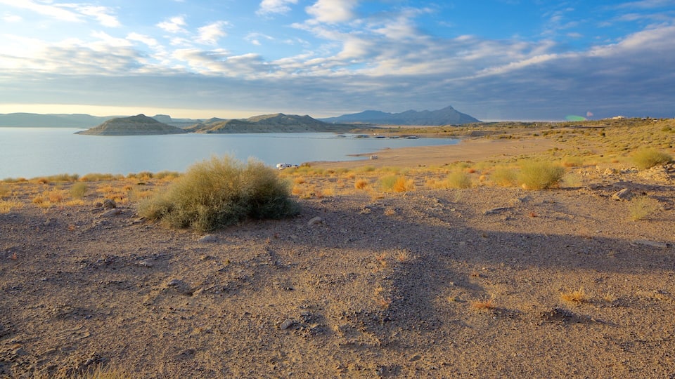 A dry, rocky landscape with sparse vegetation, a body of water, and distant mountains under a partly cloudy sky.