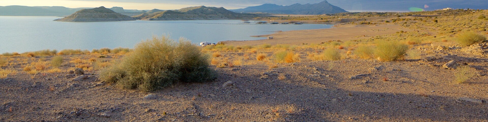 A dry, rocky landscape with sparse vegetation, a body of water, and distant mountains under a partly cloudy sky.
