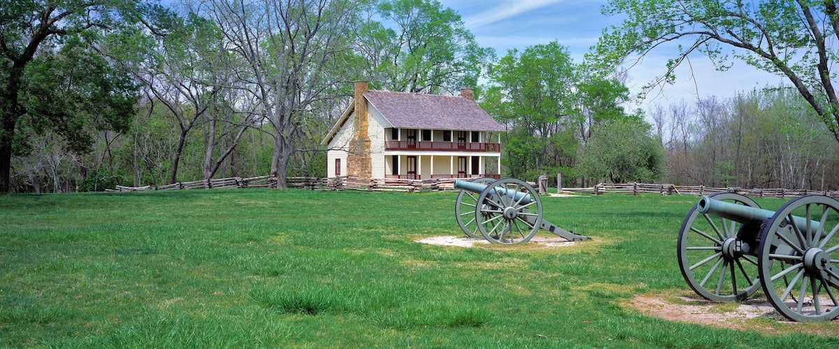 A historic house with a porch, surrounded by a wooden fence, and two cannons in a grassy field.