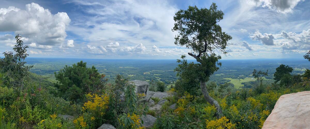 A mountainous landscape with dense greenery, rocky outcrops, and a distant view of a valley under a partly cloudy sky.
