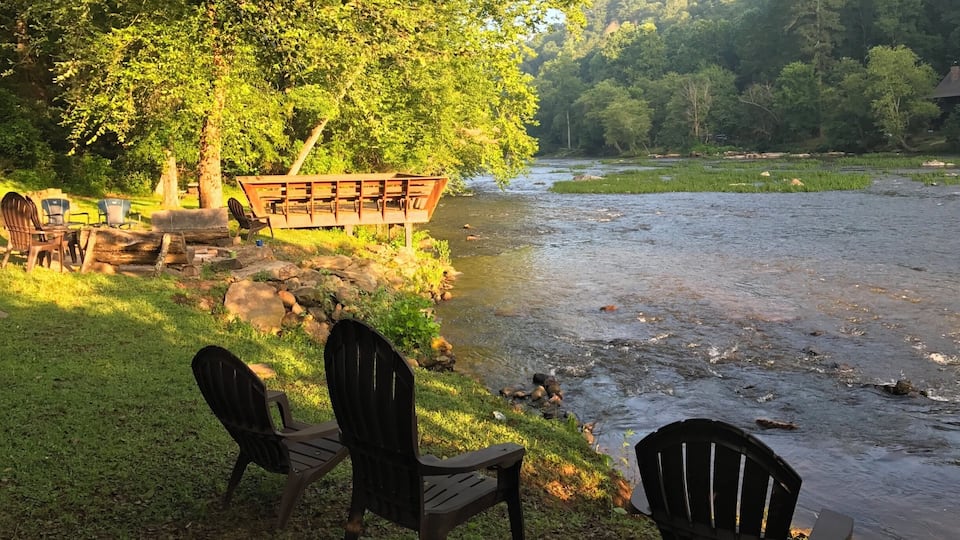 A riverside picnic area with chairs and a wooden bench overlooking a flowing river, surrounded by lush greenery and hills.