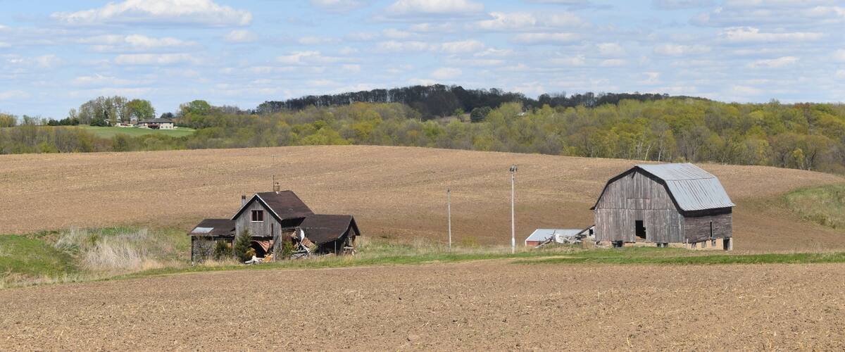 A rural landscape with two barns, a plowed field, and a forest in the background.