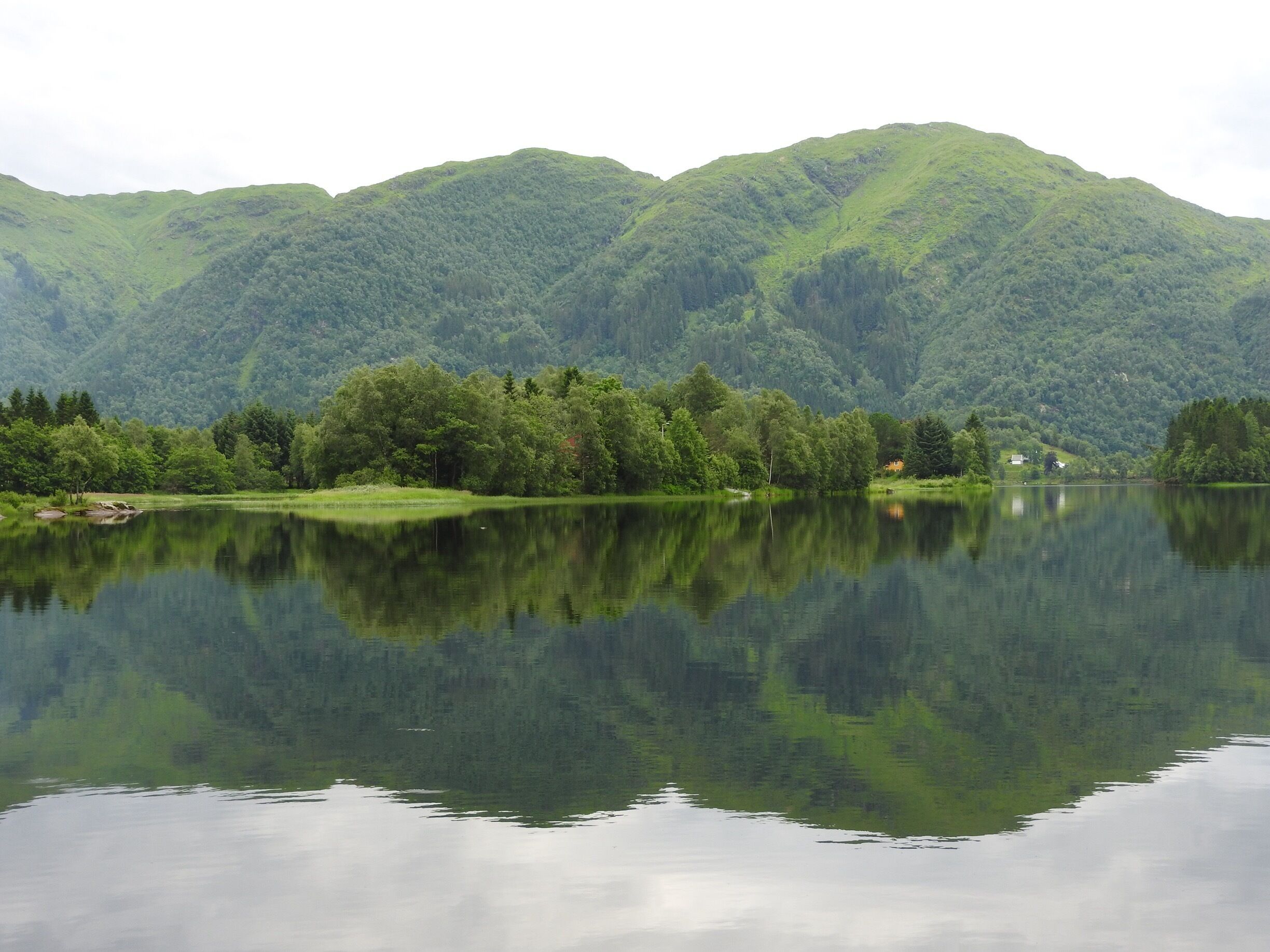 A calm lake reflecting green mountains and a small house on the shore.