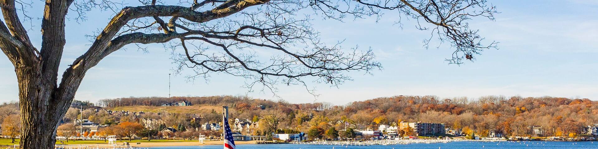 A deck with a table, chairs and an American flag above a lake on a wooded hill overlooking a town in the autumn.