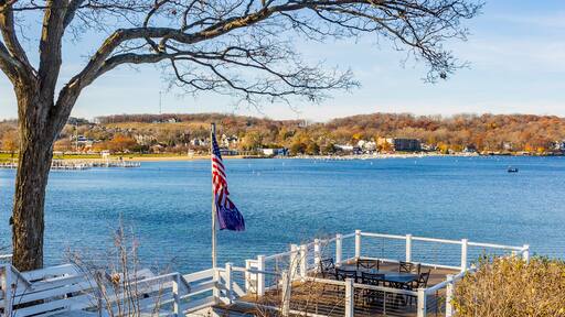 A deck with a table, chairs and an American flag above a lake on a wooded hill overlooking a town in the autumn.