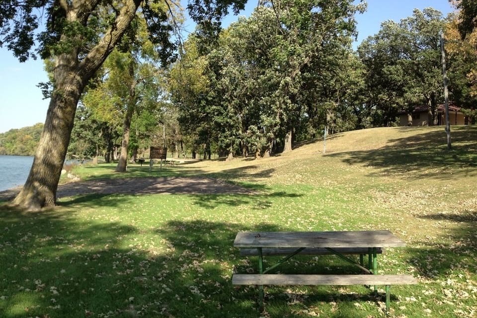 A park with a grassy area, trees, a bench, and a lake in the background.
