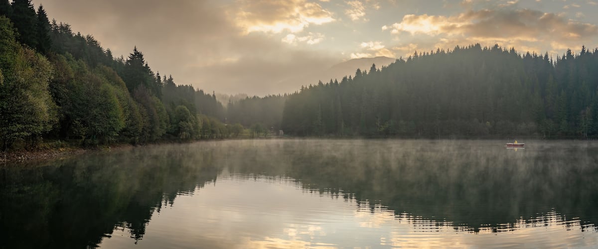 A calm lake surrounded by dense forest and misty mountains.