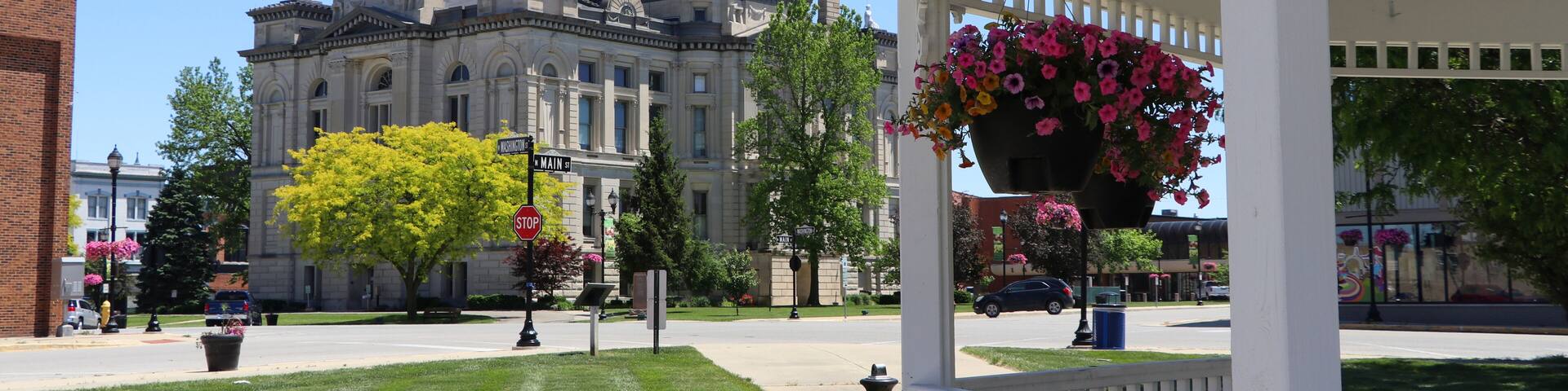 A gazebo with a hanging flower basket, a picnic table, and a historic building with a clock tower in the background.