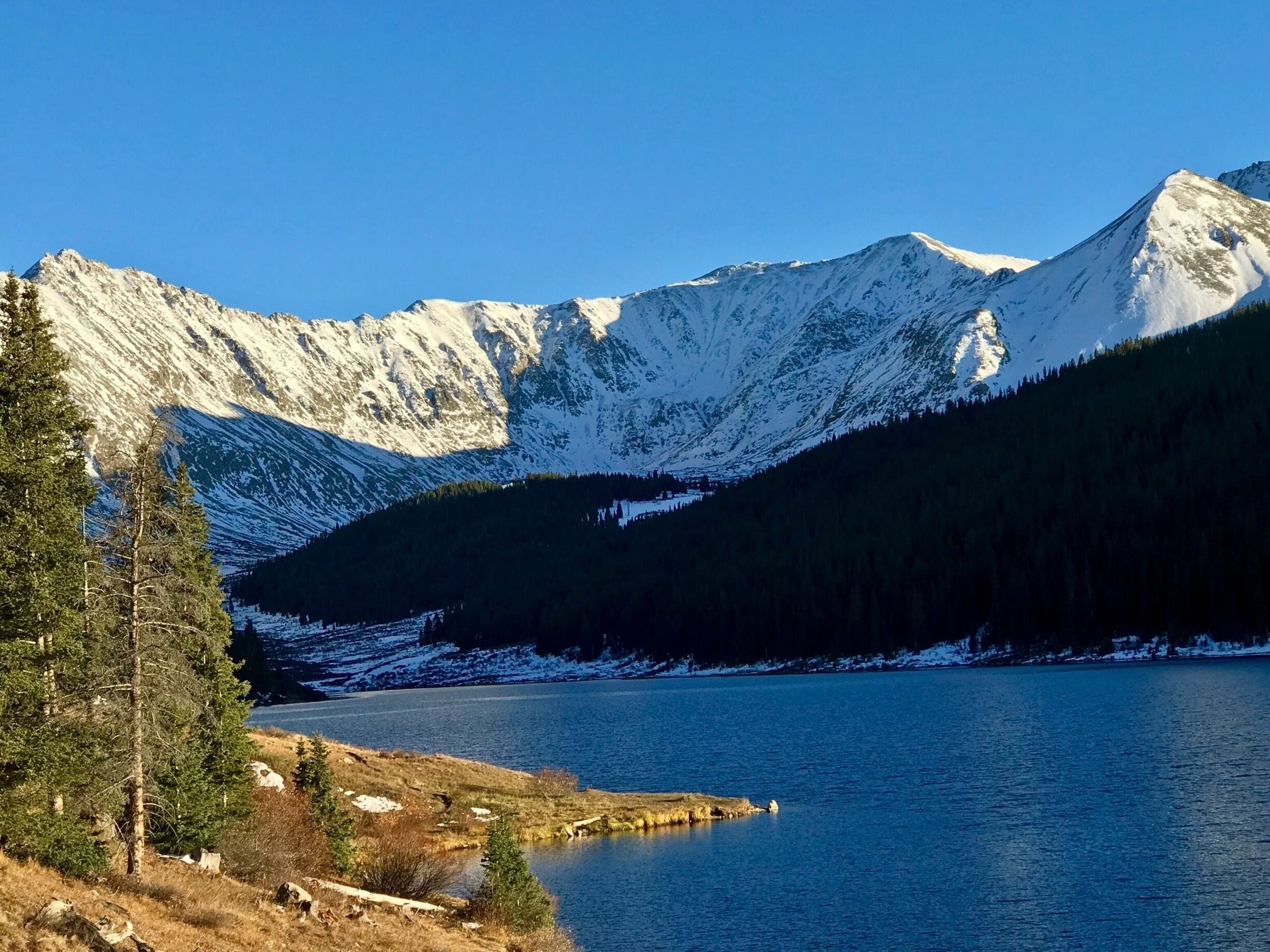 A mountain range with snow-covered peaks, a forested valley, and a calm lake.