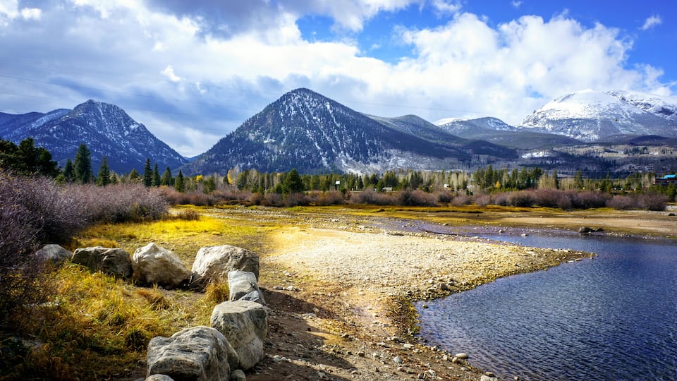 A river with a rocky shoreline, surrounded by mountains and a mix of coniferous and deciduous trees.