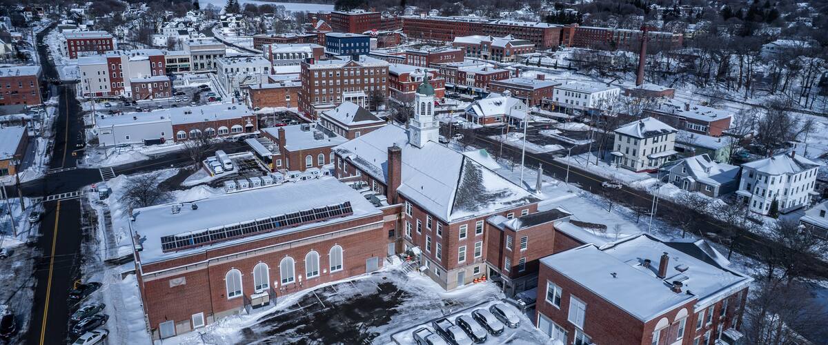 A snow-covered town with historic buildings, a river, and wind turbines in the background.