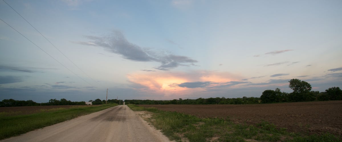 A rural road with fields on either side and a sunset sky.